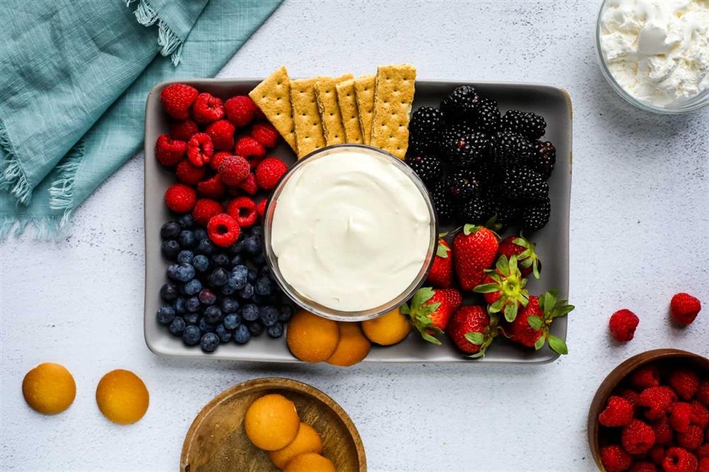 A bowl of cream in the middle of the tray surrounded by various fruits and crackers